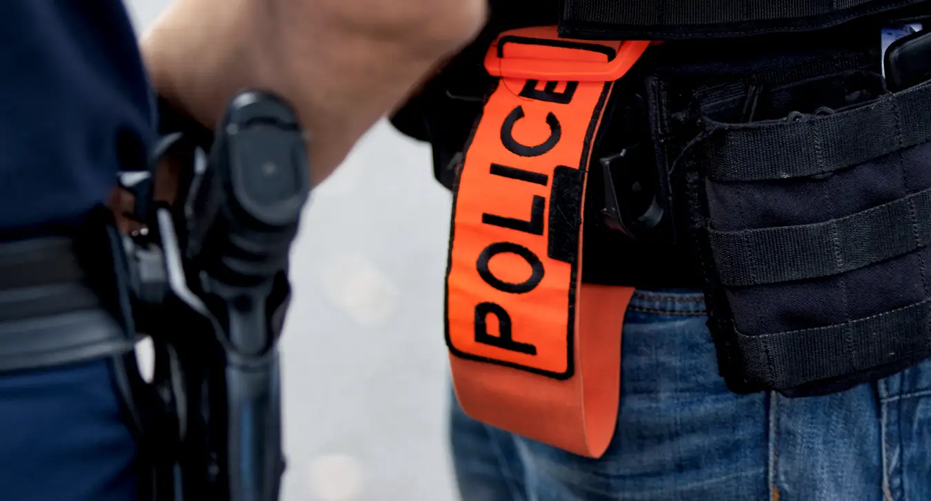 A close up of two police officers' belts. One is carrying a pistol, the other is carrying a band with the insignia POLICE