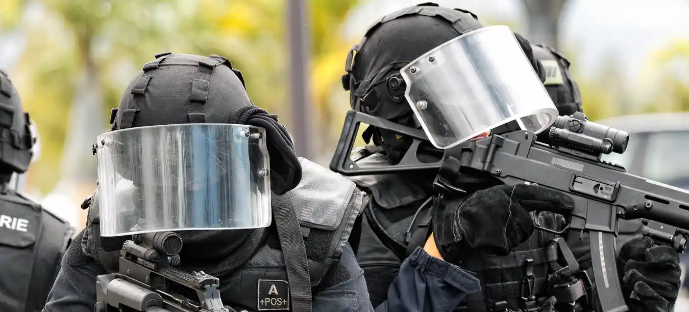Two police officers wearing heavy armour, ballistic visors and submachine guns standing ready outside