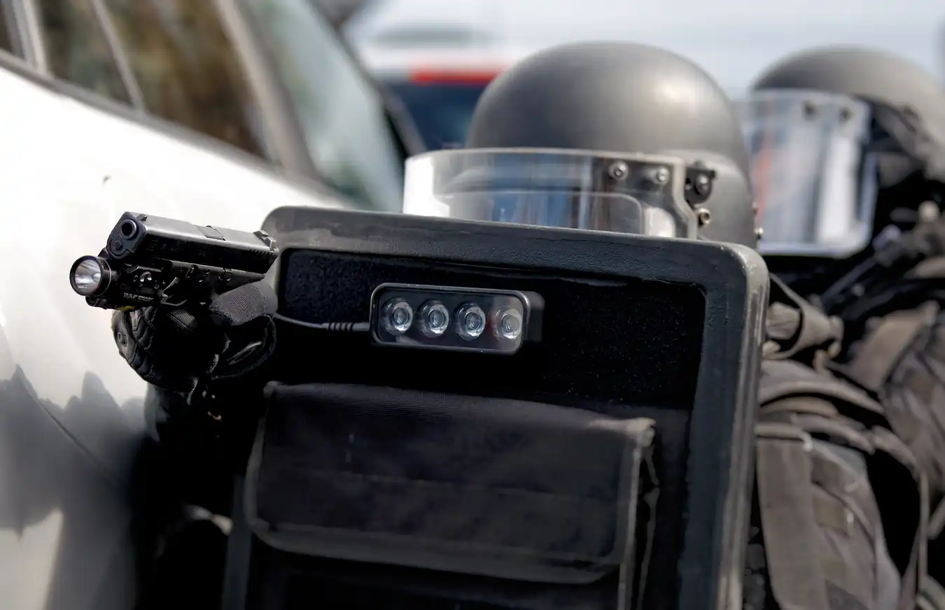 A closeup of a heavily armed police officer carrying a ballistic shield and holding out a pistol in front of a car in a police raid